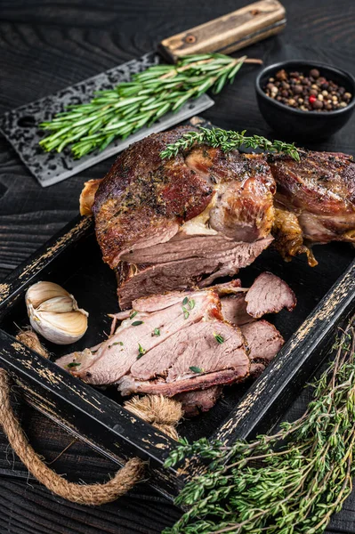 Ahşap bir tepside et satırıyla dilimlenmiş kızarmış koyun eti bacağı. Siyah ahşap arka plan. Üst görünüm