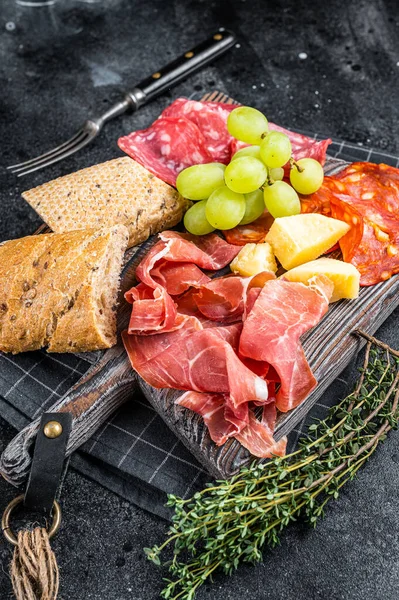 Kalte Fleischplatte, Wurst - traditionelle spanische Tapas auf einem Holzbrett mit Brot und Trauben. Schwarzer Hintergrund. Ansicht von oben — Stockfoto