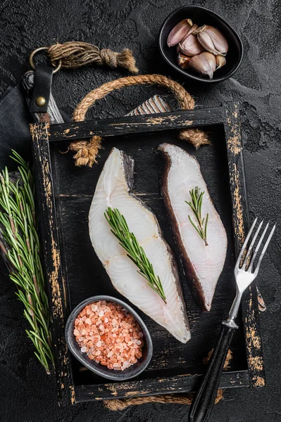 Raw halibut fish steak in wooden tray with herbs. Black background. Top view — Foto Stock