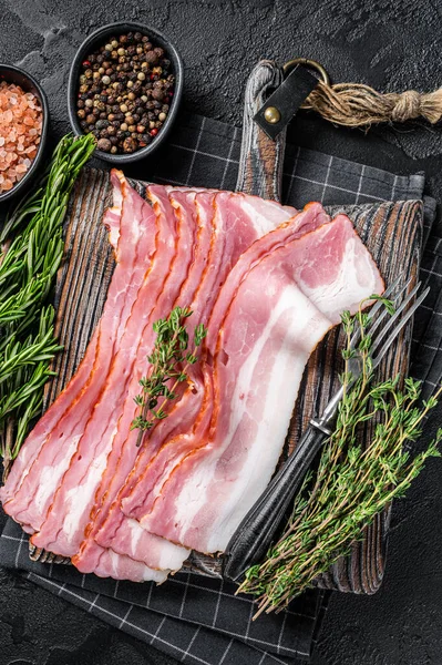 Raw smoked bacon slices with rosemary and thyme on wooden board. Black background. Top view — Stock Photo, Image