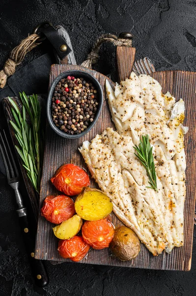 Baked Haddock fish fillet on wooden board with tomato and potato. Black background. Top view — Stockfoto