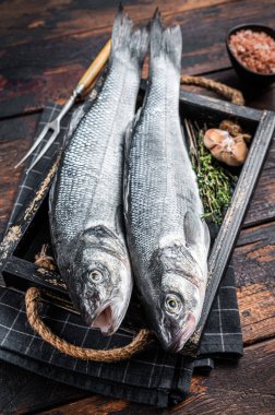 Tahta tepside otlu deniz levreği ya da çiğ balık. Koyu ahşap arka plan. Üst görünüm