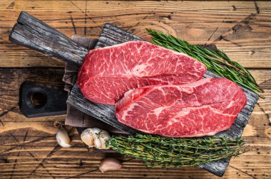 Tahta bir kasap tahtasında çiğ omuz üstü bıçaklı biftek ve otlar. Ahşap arka plan. Üst Görünüm