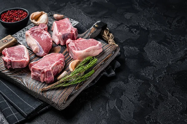 Fresh Raw lamb loin chops steaks, cutlets on butcher board with meat cleaver. Black background. Top view. Copy space