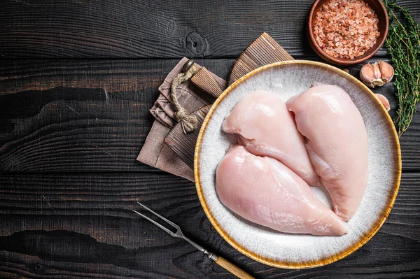 Chicken breast fillet raw meat in plate with herbs. Black Wooden background. Top view. Copy space