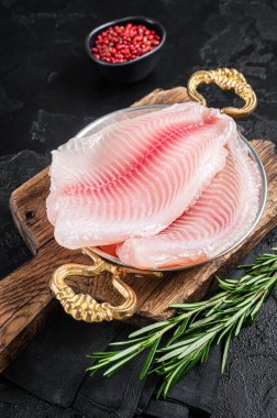 Bitkilerle dolu bir tavada çiğ tilapia balığı filetosu pişirmeye hazırım. Siyah arka plan. Üst görünüm