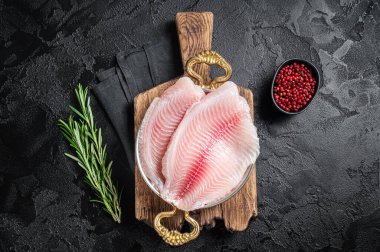 Bitkilerle dolu bir tavada çiğ tilapia balığı filetosu pişirmeye hazırım. Siyah arka plan. Üst görünüm