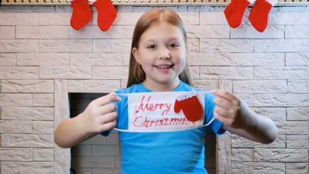 在大流行病中庆祝圣诞节 金发碧眼的小女孩戴上白色的面具 上面写着快乐的圣诞佳节 竖起大拇指 — 图库视频影像