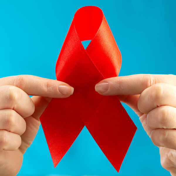 Health care and medicine concept. AIDS awareness. Female hands holding red AIDS awareness ribbon on blue background, square photo. International AIDS Day