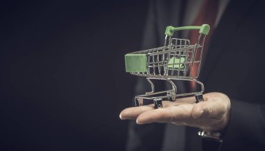 İş ve e-ticaret kavramı. Siyah arka planda, metin için fotokopi alanı olan küçük bir alışveriş arabası gösteren bir işadamı.
