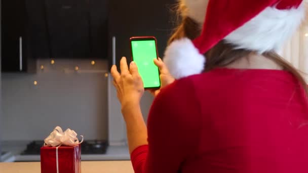 指で緑の画面を持つスマートフォンを介して台所の葉の中の新年の赤いスーツの女の子 — ストック動画