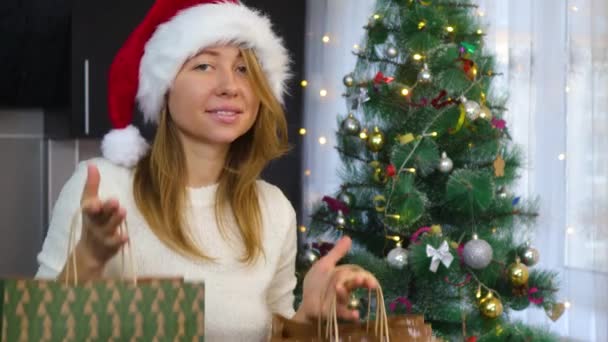 En flicka i en röd mössa dansar med paket med gåvor på bakgrunden av en julgran — Stockvideo