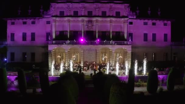La gente celebra por la noche en la villa italiana con retroiluminación púrpura, fuentes pirotécnicas — Vídeo de stock