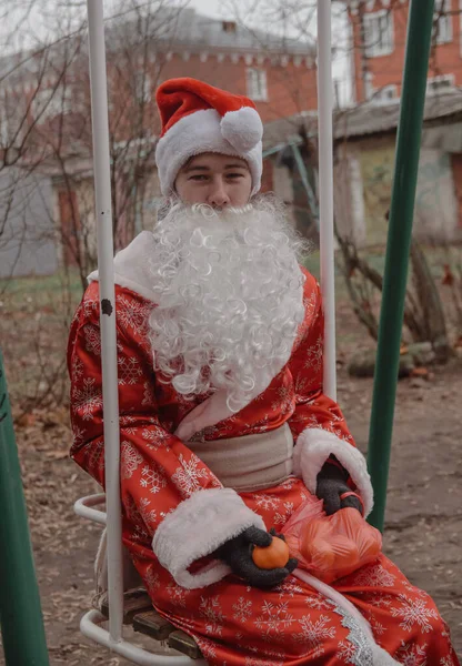 一个人穿着圣诞老人的衣服 包里夹着橘子 坐在车库后面的儿童秋千上 冬季没有雪的气候变暖的环境问题 — 图库照片