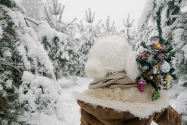 Koyun derisi ceketli bir kız, eldivenler, kışın karlı bir ormanda kulaklık takan beyaz bir şapka omzunda küçük yapay bir Noel ağacı ile huş ağacının arkasında duruyor.