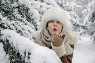 Koyun derisi ceketli, eldivenli gülümseyen genç Avrupalı bir kızın yatay kış portresi. Kulaklık ve çizmeli beyaz bir şapka. Karla kaplı çam ağaçlarında duruyor ve eldivenlerden kar üflüyor.