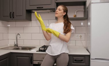 Genç esmer kız, temizlik yapmadan önce gri ve beyaz bir mutfakta dururken lastik sarı eldivenler giyer.