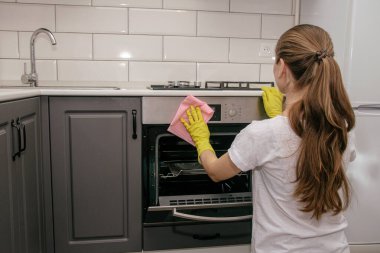 Lastik sarısı eldivenli genç bir esmer kız gri ve beyaz bir mutfakta fırını bez parçasıyla ovuyor. Ev temizliği.