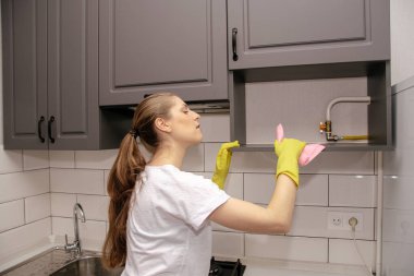 Lastik sarısı eldivenli genç esmer kız gri ve beyaz bir mutfaktaki dolaptan bir paçavrayı bir rafa sürüyor.