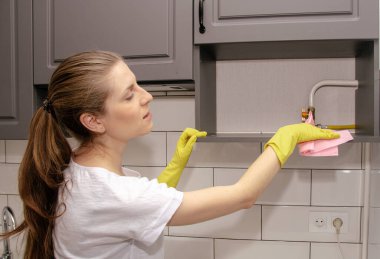 Lastik sarısı eldivenli genç esmer kız gri ve beyaz bir mutfaktaki dolaptan bir paçavrayı bir rafa sürüyor.