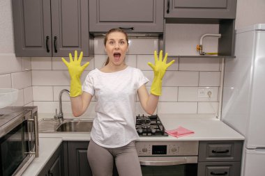 Kauçuk sarısı eldivenli genç esmer kız beyaz tişörtlü, gri taytlı gri bir mutfakta şoktan ağzı açık kalmış.