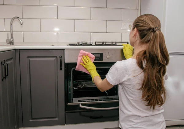 Lastik eldivenli esmer kız fırını gri ve beyaz bir mutfakla ovuyor.
