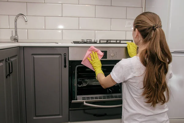 Lastik sarısı eldivenli genç bir esmer kız gri ve beyaz bir mutfakta fırını bez parçasıyla ovuyor. Ev temizliği.