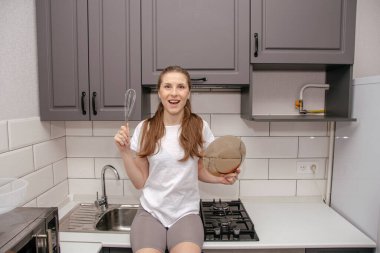 Genç esmer kız gri ve beyaz bir mutfakta çırpıcıyla ve tabağıyla oynaşıyor, gülümsüyor ve onları elinde tutuyor.