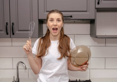 Genç esmer kız gri ve beyaz bir mutfakta çırpıcıyla ve tabağıyla oynaşıyor, gülümsüyor ve onları elinde tutuyor.