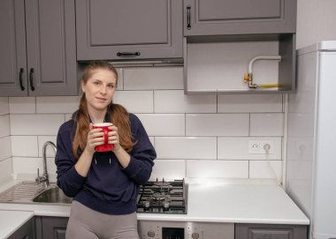 Genç bir esmer kız sabah mutfakta tek başına kahvaltı için duruyor ve yüzünde bir gülümsemeyle kırmızı bardaktan su içiyor.