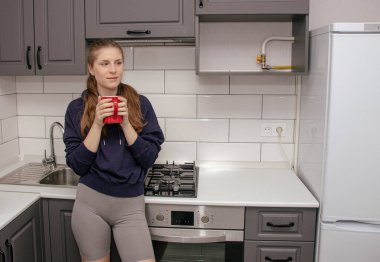 Genç bir esmer kız sabah mutfakta tek başına kahvaltı için duruyor ve yüzünde bir gülümsemeyle kırmızı bardaktan su içiyor.