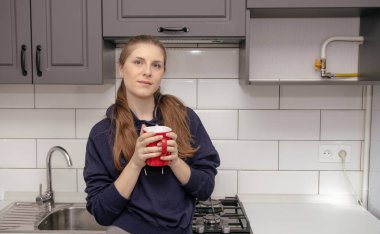 Genç bir esmer kız sabah mutfakta tek başına kahvaltı için duruyor ve yüzünde bir gülümsemeyle kırmızı bardaktan su içiyor.