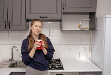 Genç bir esmer kız sabah mutfakta tek başına kahvaltı için duruyor ve yüzünde bir gülümsemeyle kırmızı bardaktan su içiyor.