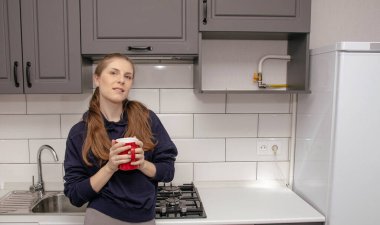 Genç bir esmer kız sabah mutfakta tek başına kahvaltı için duruyor ve yüzünde bir gülümsemeyle kırmızı bardaktan su içiyor.