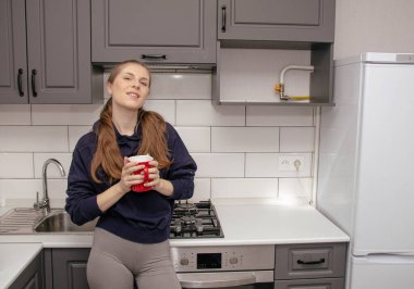 Genç bir esmer kız sabah mutfakta tek başına kahvaltı için duruyor ve yüzünde bir gülümsemeyle kırmızı bardaktan su içiyor.