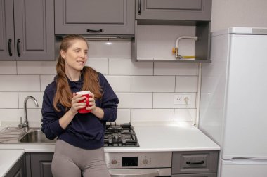 Genç bir esmer kız sabah mutfakta tek başına kahvaltı için duruyor ve yüzünde bir gülümsemeyle kırmızı bardaktan su içiyor.