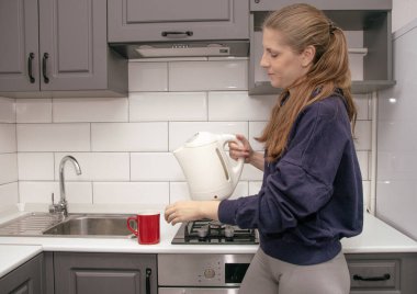 Genç, esmer bir kız kahvaltı için mutfakta tek başına durur ve çay fincanına elektrikli çaydanlıktan su döker.