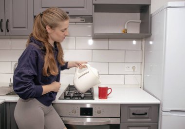 Genç, esmer bir kız kahvaltı için mutfakta tek başına durur ve çay fincanına elektrikli çaydanlıktan su döker.