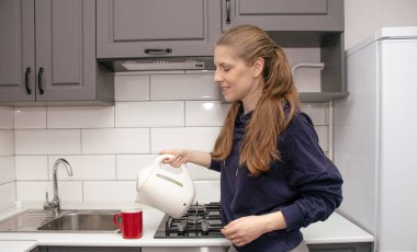 Genç, esmer bir kız kahvaltı için mutfakta tek başına durur ve çay fincanına elektrikli çaydanlıktan su döker.