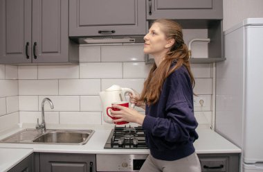 Genç, esmer bir kız kahvaltı için mutfakta tek başına durur ve çay fincanına elektrikli çaydanlıktan su döker.