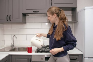 Genç, esmer bir kız kahvaltı için mutfakta tek başına durur ve çay fincanına elektrikli çaydanlıktan su döker.