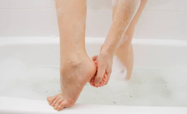 The girl in the bathroom in the foam rubs the rough heels of her feet with a washcloth