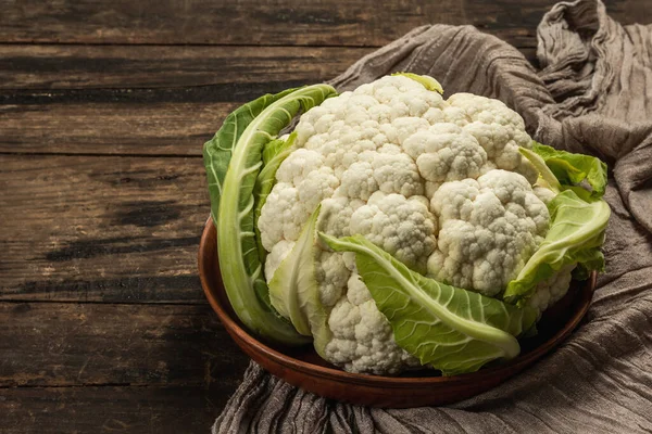 Beautiful cauliflower with green leaves in a ceramic bowl. Vintage napkin, old wooden table, copy space