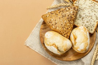 Buğday kulaklı, çörekli ve tostlu taze pişmiş ekmek. Açık bej arka planda çeşitli fırınlar. Eski keten peçete, ahşap tabak, üst manzara.