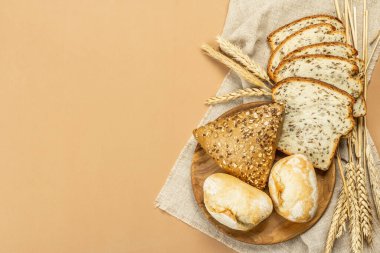 Buğday kulaklı, çörekli ve tostlu taze pişmiş ekmek. Açık bej arka planda çeşitli fırınlar. Eski keten peçete, ahşap tabak, üst manzara.