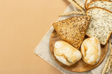 Buğday kulaklı, çörekli ve tostlu taze pişmiş ekmek. Açık bej arka planda çeşitli fırınlar. Eski keten peçete, ahşap tabak, üst manzara.