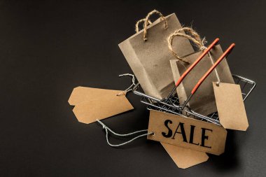 Kara Cuma indirimi konsepti. Alışveriş sepeti, el yapımı etiketler, kağıt torbalar. Siyah taş zemin, modaya uygun sert ışık, karanlık gölge, kopyalama alanı