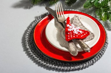 Noel sofrası. Klasik çatal bıçaklı kırmızı seramik tabak, köknar ağacı dalları ve şenlikli aksesuarlar. Modern sert ışık, karanlık gölge. Yaratıcı düz döşeme, gri arkaplan, kapat