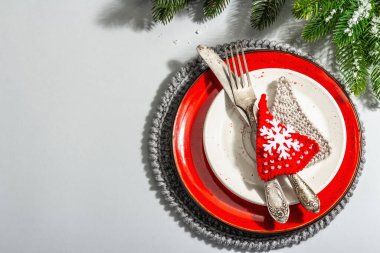 Noel sofrası. Klasik çatal bıçaklı kırmızı seramik tabak, köknar ağacı dalları ve şenlikli aksesuarlar. Modern sert ışık, karanlık gölge. Yaratıcı düz konum, gri arkaplan, üst görünüm