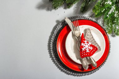 Noel sofrası. Klasik çatal bıçaklı kırmızı seramik tabak, köknar ağacı dalları ve şenlikli aksesuarlar. Modern sert ışık, karanlık gölge. Yaratıcı düz konum, gri arkaplan, üst görünüm